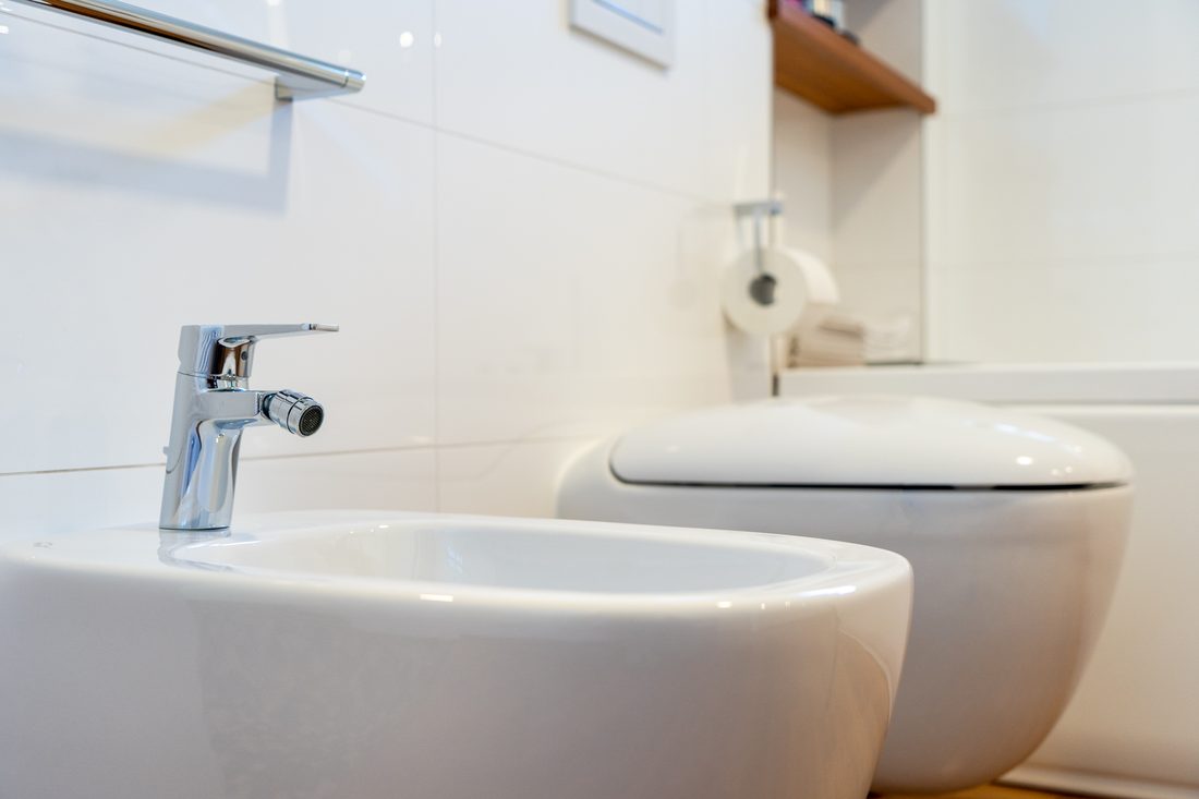 Detail bidetu s chromovou baterií, za ním závěsné WC a bílé velkoformátové obklady